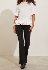 Bílé volánkové tričko s krátkými rukávy a volánkovým lemem, kombinované s černými rozšířenými džíny a kotníkovými botami, na neutrálním pozadí.