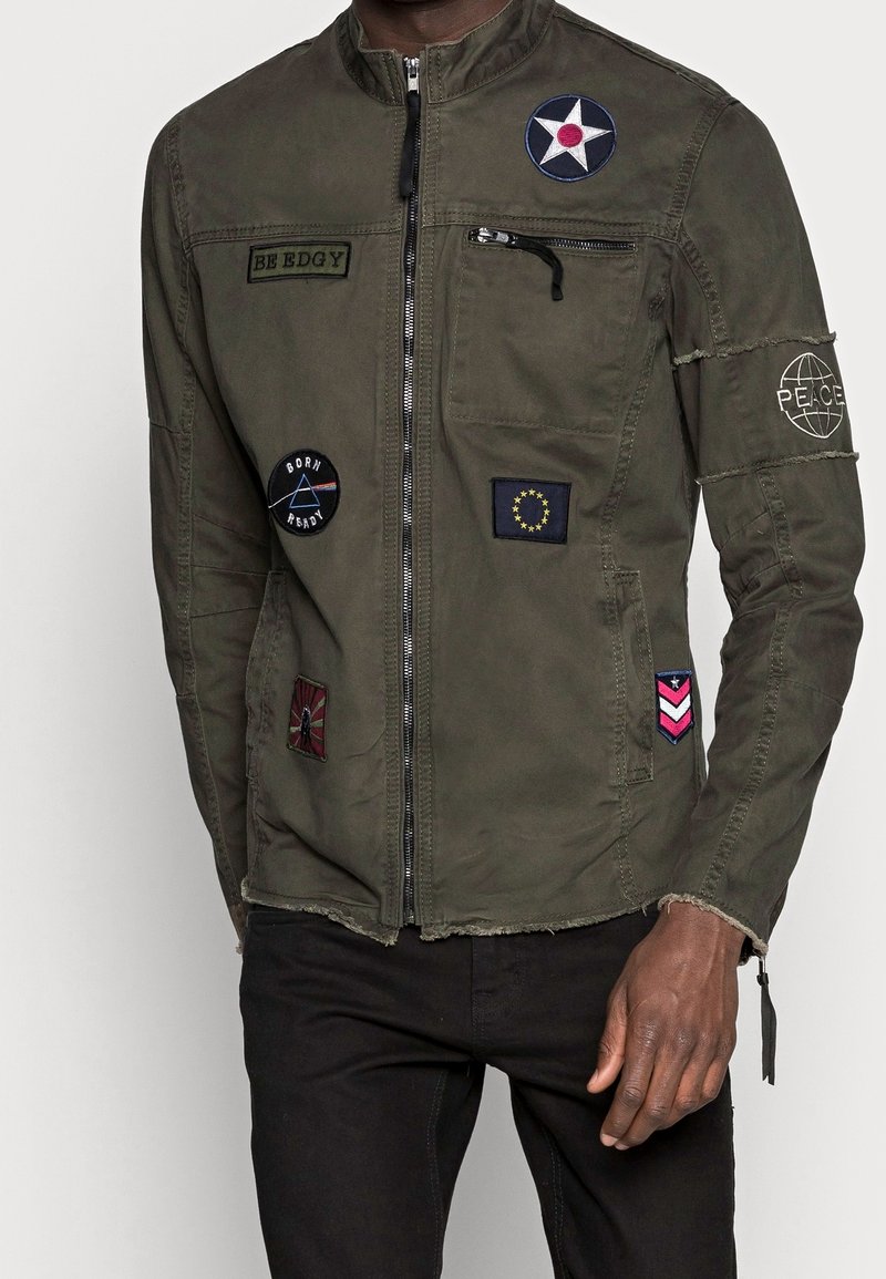 Grüne Militärjacke mit Stehkragen, durchgehendem Frontreißverschluss und verschiedenen aufgestickten Abzeichen in unterschiedlichen Farben und Designs.