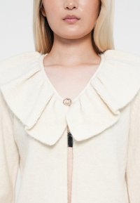Kremfarget cardigan med en stor, ribbet, brettet krage og en dekorativ perleknapp ved halsen. Myk tekstur, åpen frontdesign.