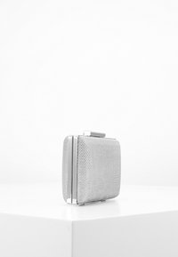 Lille sølvfarvet clutch-taske med struktureret overflade, metalkanter og lås, placeret på en hvid overflade mod en ensfarvet hvid baggrund.