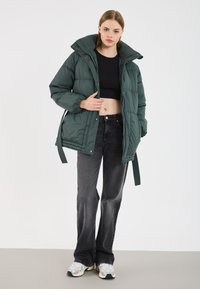Donkergroene pufferjack met een hoge kraag, voorzien van een ritssluiting en knoopsluiting, gecombineerd met zwarte flared jeans en sneakers.