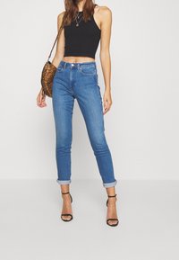 Högmidjade blå jeans i denim med en figursydd design, rullade fållar och en mjuk textur, kombinerade med en svart crop top och en mönstrad handväska.