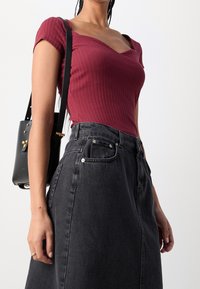 Femme portant un haut côtelé bordeaux, une jupe en jean noire taille haute et un sac à épaule noir avec des détails dorés sur un fond uni.