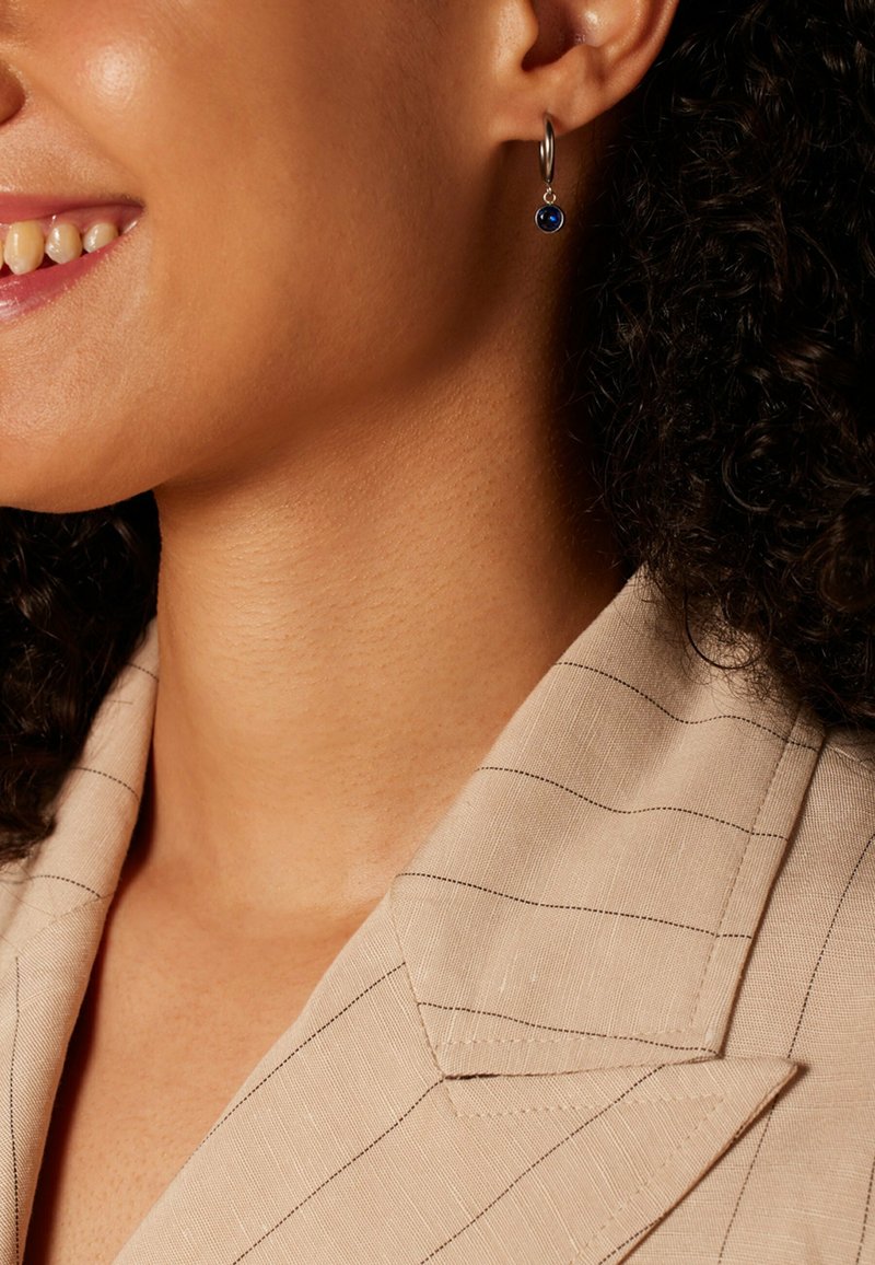 Zilveren créole met een blauwe edelsteen, met een gladde oppervlakte en een subtiele glans, gedragen door een model met krullend haar en een gestreepte blouse.