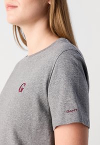 Pilka medvilninė marškinėlių su apvalia iškirpte. Mažas bordo spalvos "G" logotipas ant krūtinės ir "GANT" siuvinys ant rankovės. Lygi tekstūra.