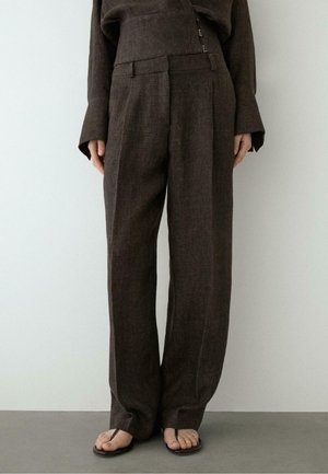 Brązowe spodnie z szerokimi nogawkami i wysokim stanem, w zestawie z dopasowanym topem z długim rękawem, noszone z czarnymi sandałami typu thong na szarej podłodze.