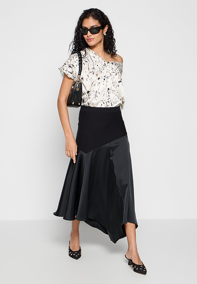 Off-shoulder witte top met zwart abstract patroon, gecombineerd met een zwarte asymmetrische satijnen rok. Accessoires omvatten kitten-heel schoenen en een tas.