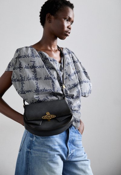 Bolsa de ombro em pele preta com formato curvado e detalhes em hardware dourado. Usada com uma blusa estampada azul e branca e jeans de denim claro.