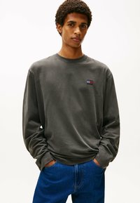 Camiseta de manga larga gris de tejido suave, con un cuello de canalé, presenta un pequeño logo multicolor en el pecho. Combinada con denim azul.