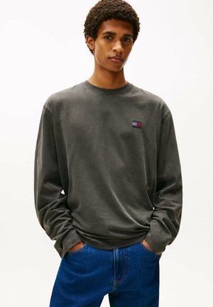 Grå langærmet skjorte i blødt stof med ribbet halsudskæring, som har et lille multikolor logo på brystet. Kombineret med blå denim.