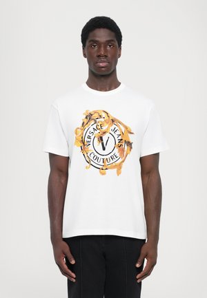 Hvit bomulls t-skjorte med et sirkulært logo-design med gull- og svart aksenter, som viser "Versace Jeans Couture" med fremtredende tekst.