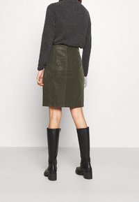 Jupe en cuir vert olive avec une texture lisse et un éclat subtil, associée à un pull en maille gris foncé et des bottes noires montantes.