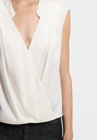 Cremefarbener, ärmelloser Blusen mit V-Ausschnitt, ausgestattet mit sanften Falten und einer glatten Textur. Kombiniert mit dunklem Denim.