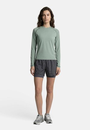 Femme debout, face à l'avant, portant une chemise à manches longues vert clair, un short gris foncé et des chaussures de sport grises sur un fond blanc.