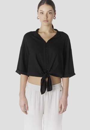 Ung kvinde iført en sort crop skjorte med knapper, bundet ved taljen, og løse hvide bukser, stående foran en ensfarvet baggrund.