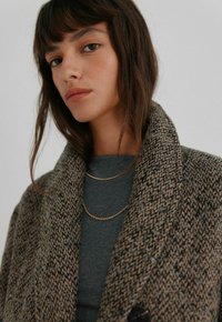 Uldblandingsfrakke med et tekstureret, brunt og sort vævet mønster, med en stor krave og knaplukning, båret over en mørkegrå top og guldkæder.