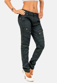 I jeans skinny verde scuro con motivo camouflage presentano un design multi-tasche con cerniere, una vestibilità aderente e un tessuto testurizzato. Sono visibili tacchi alti bianchi.