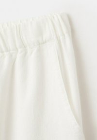 Gros plan sur un pantalon en tissu blanc avec une ceinture élastique et une poche latérale sur un fond uni.