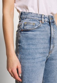 Dettaglio di una persona che indossa jeans in denim blu chiaro a vita alta e una camicia bianca infilata, con la mano sinistra rilassata al lato.