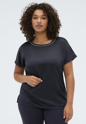 Mujer con el cabello rizado que lleva una blusa azul marino de manga corta con ribete metálico en el cuello, de pie frente a un fondo claro y liso.