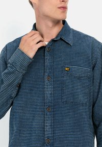 Blauw gestreepte overhemden met een puntige kraag, borstzak en bruine knopen; de stof heeft een gestructureerde afwerking. Klein logo op de zak.