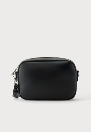 Petit sac bandoulière noir rectangulaire à texture lisse, fermeture zippée sur le dessus et sangle amovible avec accessoires argentés, sur fond uni.