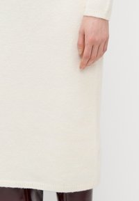 Knelang krem strikket kjole med en myk tekstur. Ribbestrikket mansjetter og høyre. En hånd hviler på stoffet nær bunnen.