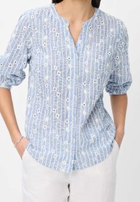 Blouse bleu clair avec motif floral blanc brodé, manches courtes bouffantes et encolure en V. Fabriquée en tissu léger et transparent.