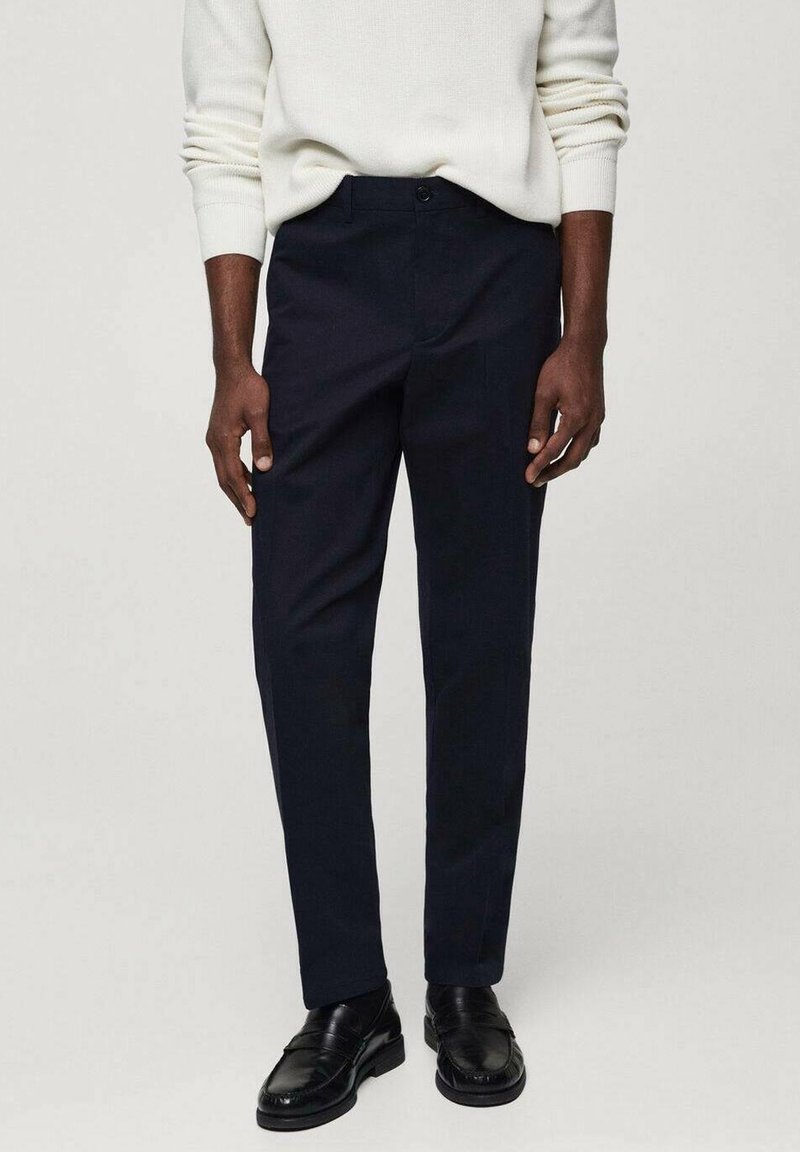 Homme portant un pantalon noir, des chaussures en cuir noires et un pull blanc à manches longues sur un fond clair uni.
