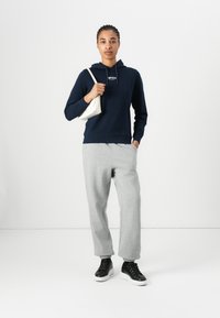 Sweatshirt à capuche marine, pantalon de survêtement gris et baskets noires. Sac à bandoulière blanc. Le modèle est debout devant un fond blanc.