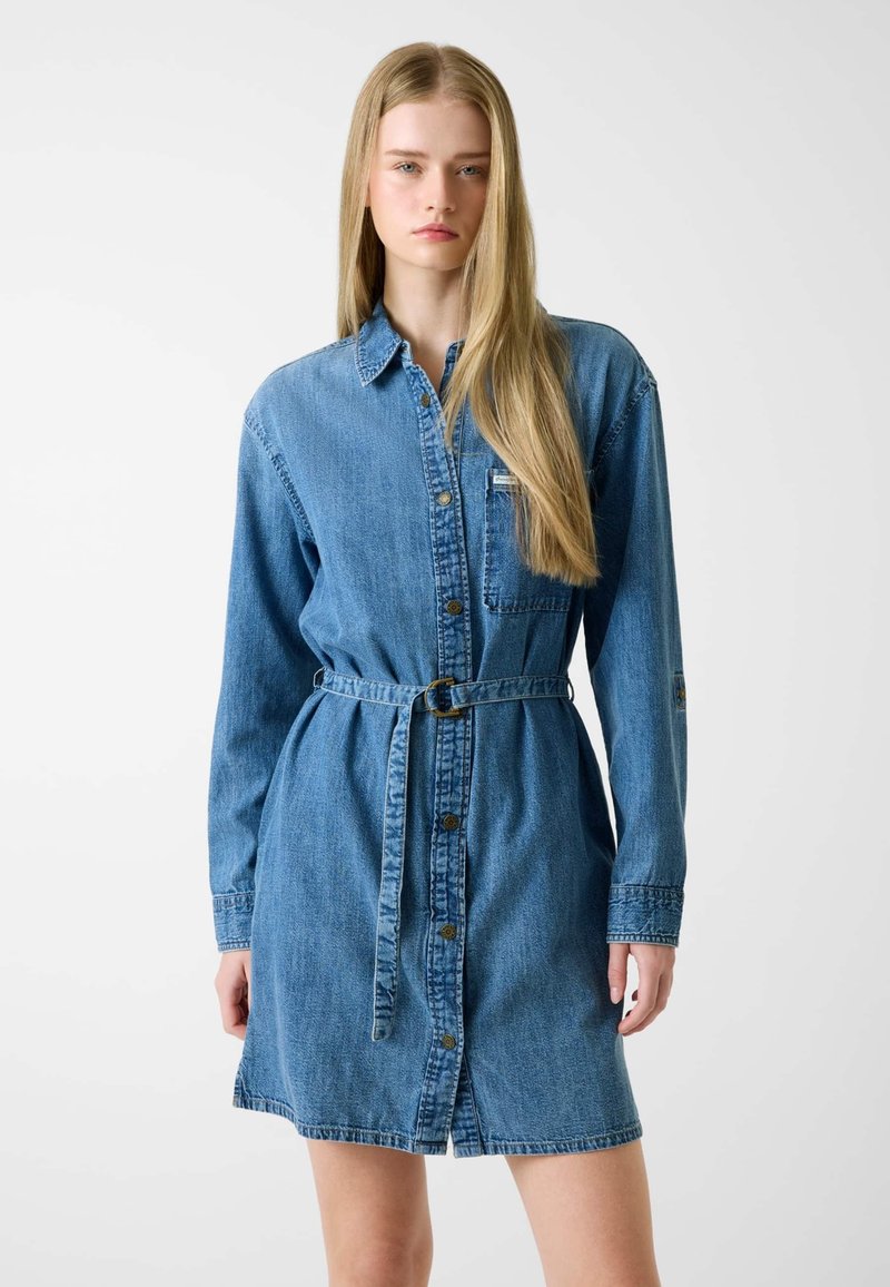 Vestito camicia in denim con chiusura frontale a bottoni, maniche lunghe, cintura in vita e una singola tasca sul petto in una tonalità medio-blu.