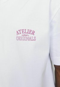 Bijela pamučna majica s ružičastim izvezenim tekstom "ATELIER" i "ORIGINALS" s lijeve strane, kratke rukave i standardni kroj.
