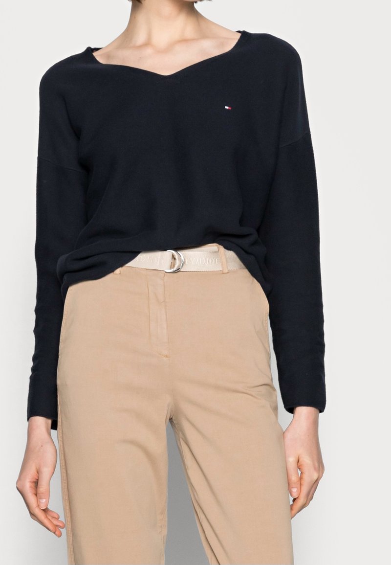 Pull en maille bleu marine avec un design à col en V, coupe légèrement ample, associé à un pantalon beige et une ceinture claire avec une boucle ronde.