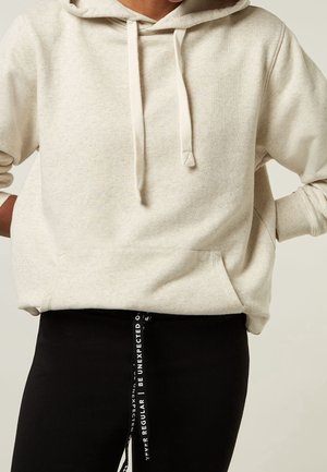 Personne portant un sweat à capuche beige clair avec cordons de serrage et un pantalon noir avec une bande verticale à texte blanc sur la ceinture.