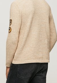 Beige gestrickter Pullover mit strukturiertem Muster, runder Ausschnitt und gerippten Bündchen. Verfügt über ein Abzeichen am linken Ärmel mit Stickerei.