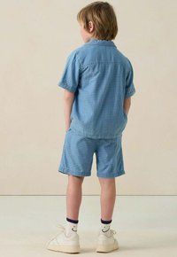 Jungenoutfit mit einem kurzärmeligen Hemd mit Knöpfen und einer kurzen Hose in Hellblau mit weißen Streifen, ergänzt durch weiße Sneakers und gestreifte Socken.