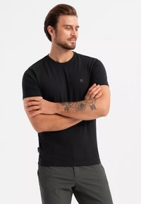 Sort kortærmet T-shirt lavet af glat bomuld. Rund halsdesign med et lille etiketmærke på siden. Modellen viser en afslappet positur.