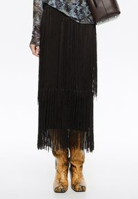 Czarna spódnica z frędzlami o fakturowanym splocie, zestawiona z brązowymi kowbojkami ze skóry węża. Strój wyróżnia się warstwowanymi wzorami i akcentami.
