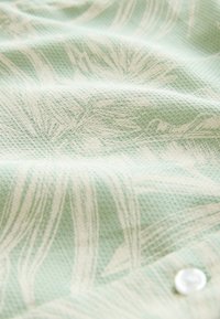 Tessuto verde chiaro con un motivo testurizzato, presenta disegni botanici in una tonalità più chiara e un dettaglio con un bottone bianco.