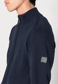 Marineblauwe zip-up trui met een hoge kraag en ribbeldetails. Heeft een leren logo-patch op de linkermouw. Gehaakt textuur over de gehele trui.