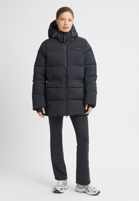 Chaqueta acolchada negra con un cuello alto, cierre de cremallera y capucha. Presenta un diseño acolchado y un detalle de logo en la parte superior izquierda.