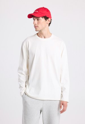 Chemise en coton crème à manches longues avec un col rond et une coupe décontractée, associée à un pantalon de survêtement gris et une casquette de baseball rouge.