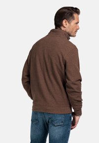 Man die zijwaarts kijkt, gekleed in een bruine gestructureerde pullover met een staande kraag en blauwe jeans, tegen een effen witte achtergrond.