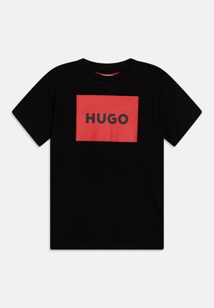 Czarna koszulka z krótkim rękawem z czerwonym prostokątnym naszywką z napisem "HUGO" pogrubionymi czarnymi literami na środku klatki piersiowej.