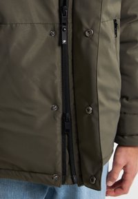 Jachetă pufosă de culoare verde măslinie, cu fermoar negru și buttoni, cu textură matlasată și buzunare laterale, expusă alături de blugi deschiși.