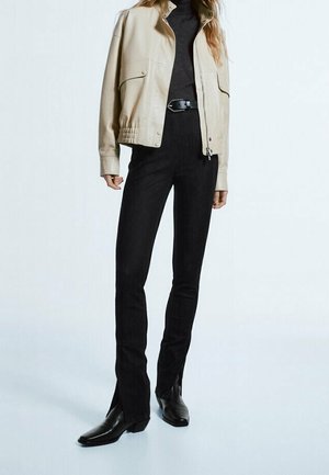 Femme portant une veste beige courte par-dessus un col roulé noir, un pantalon taille haute noir ceinturé et des bottines à talons noires sur un fond clair.