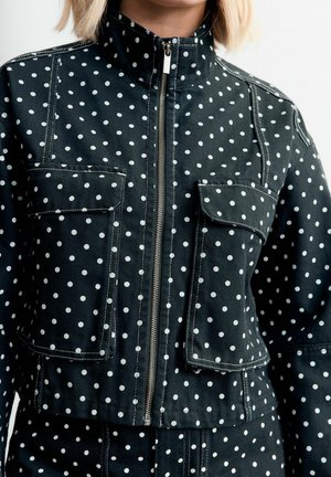 Schwarze Jacke mit Reißverschluss und weißen Punkten, zwei großen Klappentaschen auf der Brust, getragen von einer Person mit kurzen blonden Haaren.