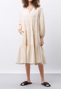 Vestido beige de capas con mangas largas y escote en V. Hecho de una tela suave con un ajuste holgado y bolsillos laterales. Combinado con zapatos negros tipo slip-on.