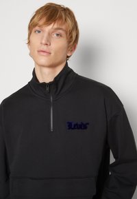 Felpa nera con zip e collo alto, caratterizzata da un design corto e un logo ricamato di un blu scuro peloso sul davanti. Texture liscia.