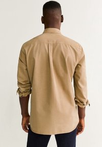 Hombre llevando una camisa de manga larga beige con las mangas arremangadas, de pie de espaldas contra un fondo claro y liso.